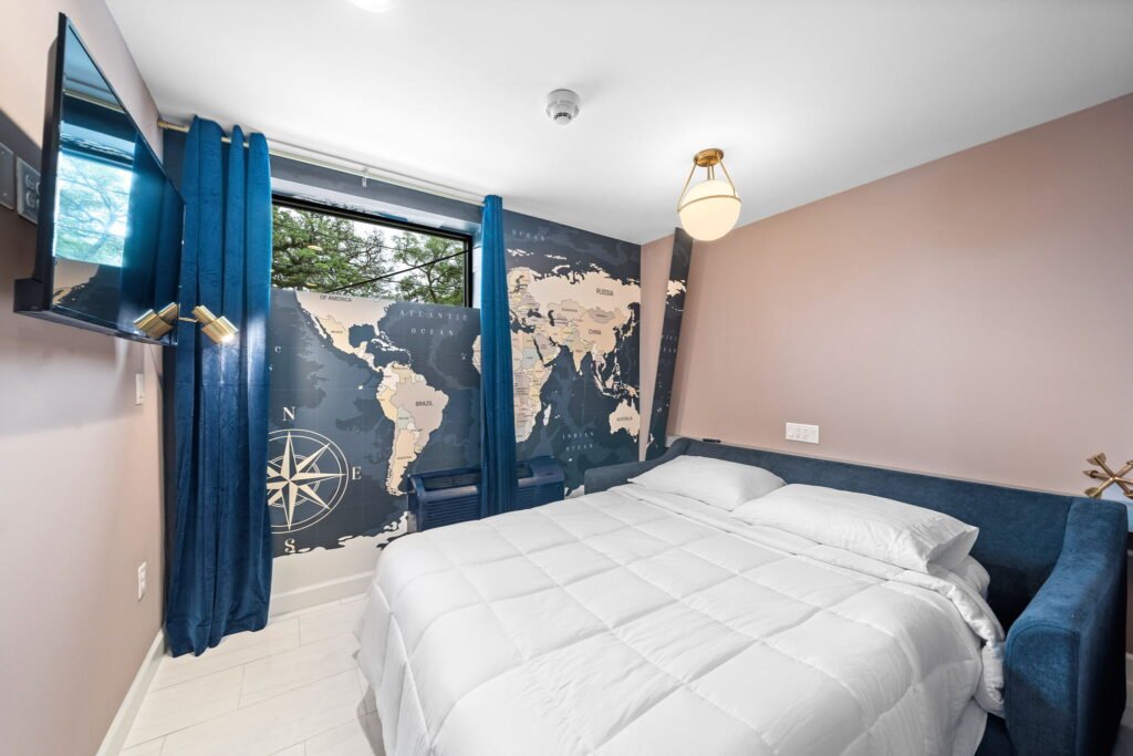 Boutique hotel room at Wanderstay featuring a queen bed with white linens, a blue accent sofa, deep blue curtains, and a large world map wall mural.
