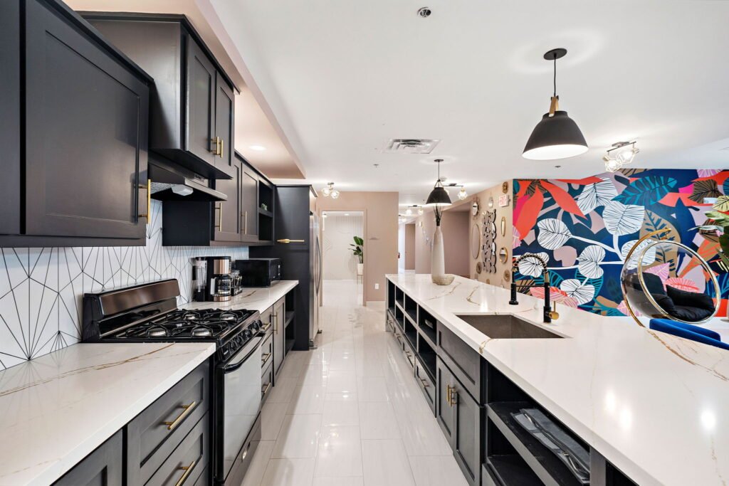 Boutique hotel shared guest kitchen featuring a long white quartz island with an integrated sink, dark gray cabinets with gold hardware, and a colorful tropical floral accent wall.